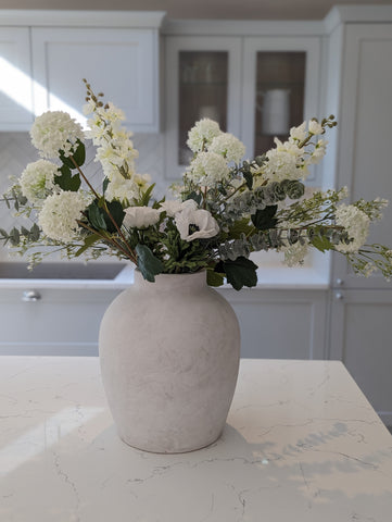 large, grey and white stone vase, on a kitchen island with a large bouquet of white and green artificial flowers