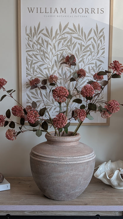 A rustic terracotta-style ceramic vase filled with pink hydrangea-style artificial flowers and green foliage, displayed on a wooden shelf in front of a framed botanical print reading William Morris.