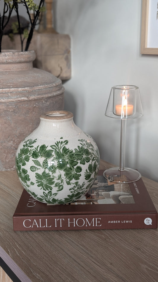 Decorative vase with green floral pattern on a book titled 'Call It Home' with a candle in the background.