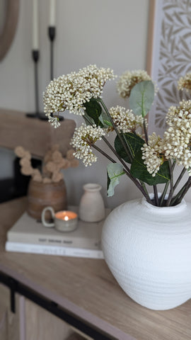 a simple white vase sits on a rustic wooden sideboard. The vase contains a beautiful arrangement of off white viburnum bud stems. a dandle burns in the badkground on a set of coffee table styling books.